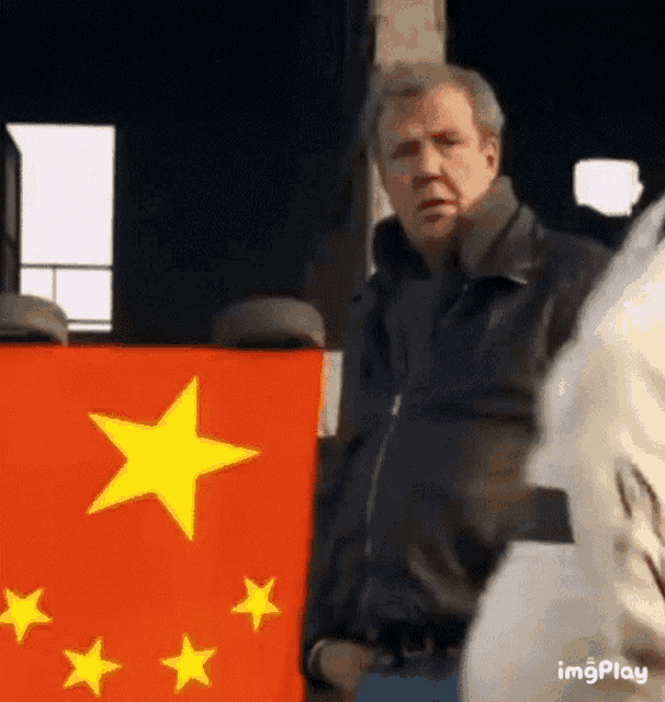 a man in a black jacket is standing next to a red and yellow flag with stars .