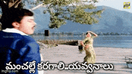 a man in a blue jacket is standing next to a woman in a green saree dancing in front of a lake .