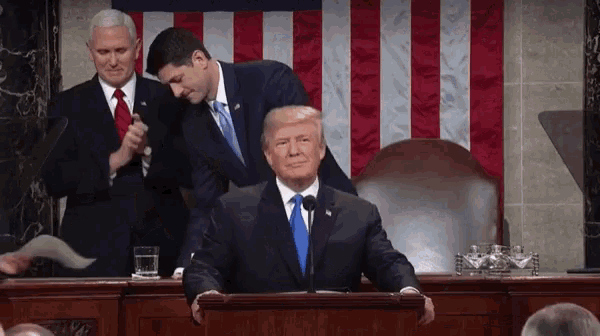 a man in a suit and tie is sitting at a podium giving a speech while two other men stand behind him .