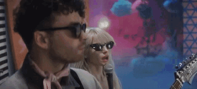 a man and a woman wearing sunglasses are standing next to each other . the man is holding a guitar .
