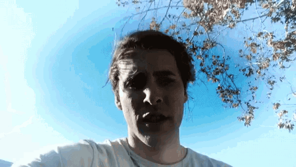 a man in a white shirt stands in front of a tree with a blue sky in the background