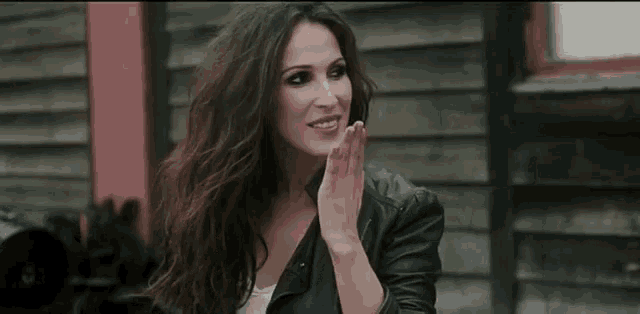 a woman wearing a leather jacket is standing in front of a wooden building and applauding .