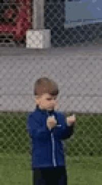 a young boy in a blue jacket is standing in front of a chain link fence on a soccer field .