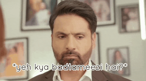 a man with a beard is standing in front of a wall of framed pictures and says " yeh kya badtameezi hai "
