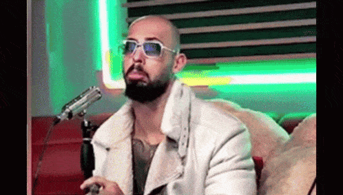 a bald man with a beard is wearing sunglasses and a white jacket while sitting in front of a microphone .