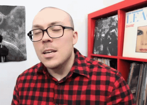 a man wearing glasses and a plaid shirt stands in front of a shelf with a lee dare album