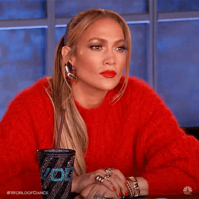 a woman wearing a red sweater is sitting at a table with a cup that says world of dance on it