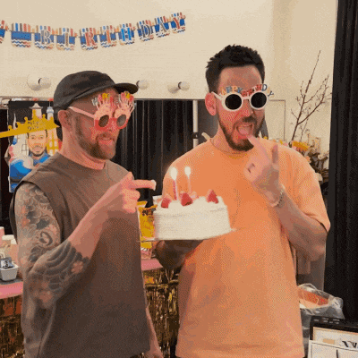 two men wearing birthday glasses holding a cake with candles