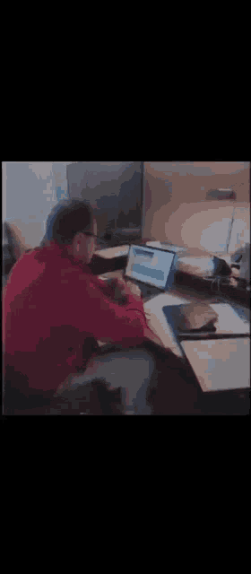a man in a red sweater is sitting at a desk with a laptop on it .