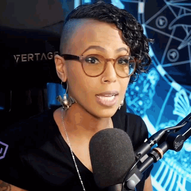 a woman wearing glasses and earrings is speaking into a microphone with a vertage chair in the background