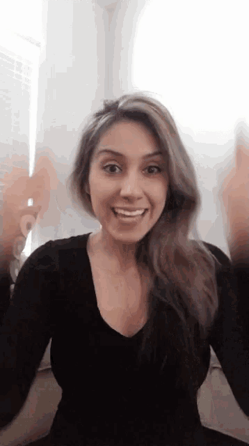 a woman in a black shirt is smiling and waving her hands in front of a window .