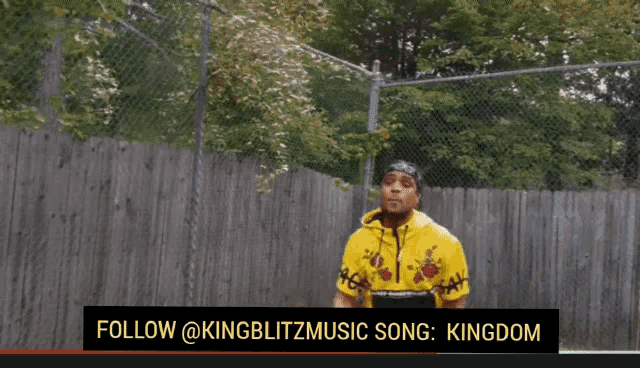 a man in a yellow hoodie is standing in front of a chain link fence with the words follow @kingblitzmusic song kingdom below him