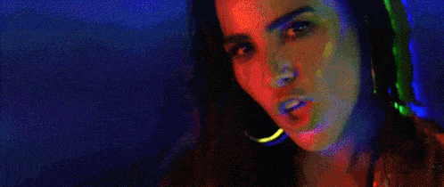 a woman is standing in front of a neon sign in a dark room and looking at the camera .
