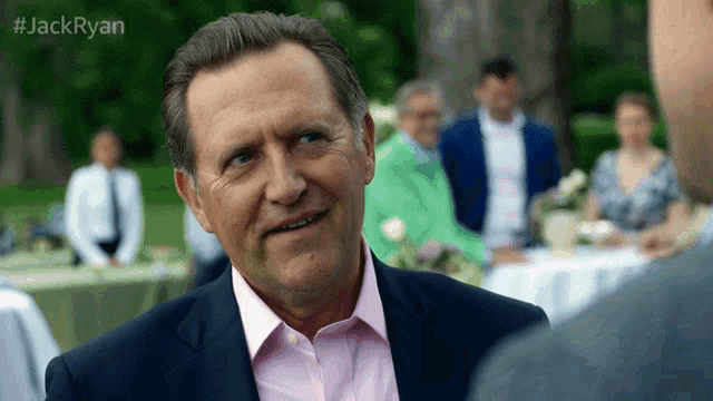 a man in a suit and pink shirt is talking to another man at a party .