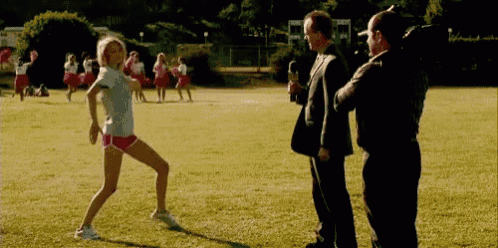 a woman in shorts is dancing in a field while a man in a suit stands behind her .