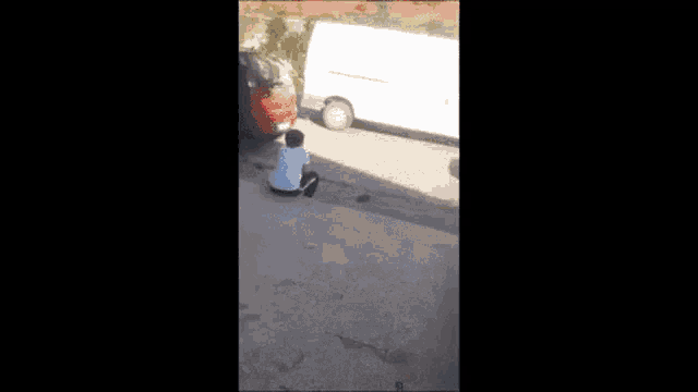 a man is sitting on a sidewalk next to a red car and a white van .