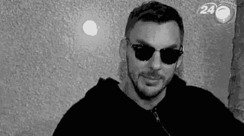 a man wearing sunglasses and a hoodie is standing in front of a wall .