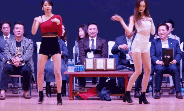 two women are dancing in front of a group of people sitting at a table with bottles of water on it