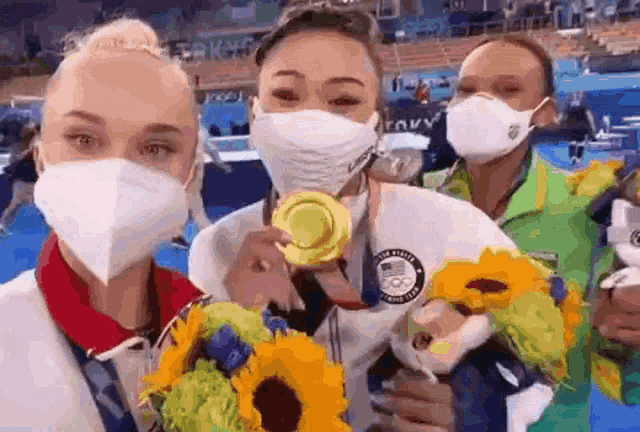 three female gymnasts wearing masks are holding flowers and medals .