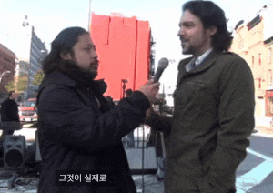 a man is talking into a microphone while another man looks on with korean writing on the bottom