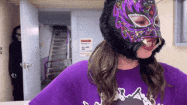 a woman wearing a purple shirt and a purple mask stands in front of stairs