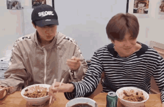 two men are sitting at a table with bowls of food and a can of soda