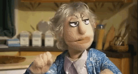 a puppet with gray hair and a mustache is sitting in a kitchen making a funny face .