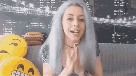 a woman with blue hair is sitting on a couch with a bunch of emoji pillows