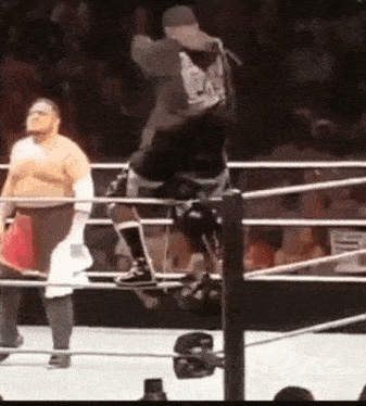 a wrestler is jumping over a rope in a wrestling ring while another wrestler watches .