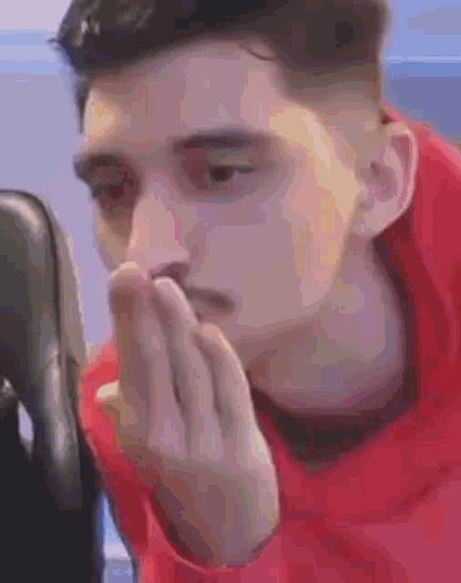 a man in a red hoodie is covering his mouth with his hands while sitting in a chair .