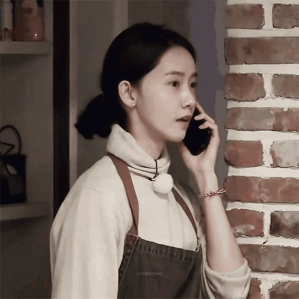 a woman in an apron is talking on a cell phone in front of a brick wall