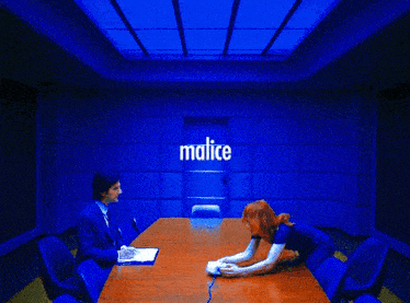 a man and a woman are sitting at a table with malice written on the wall behind them