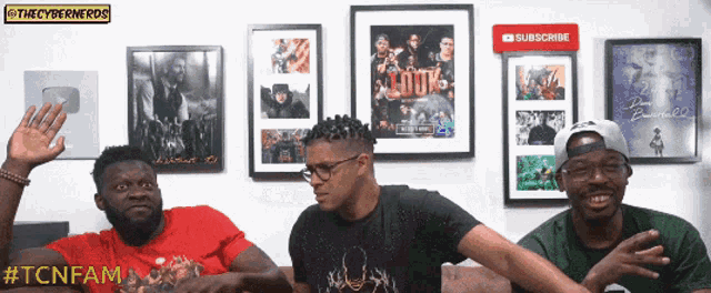 three men are sitting in front of a wall with framed pictures and a subscribe button