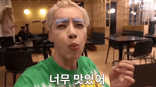 a man with blonde hair and blue eyebrows is wearing a green shirt and making a funny face in a restaurant .