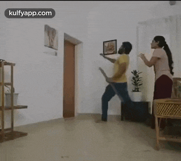 a man in a yellow shirt is standing on one leg in a room holding a broom .