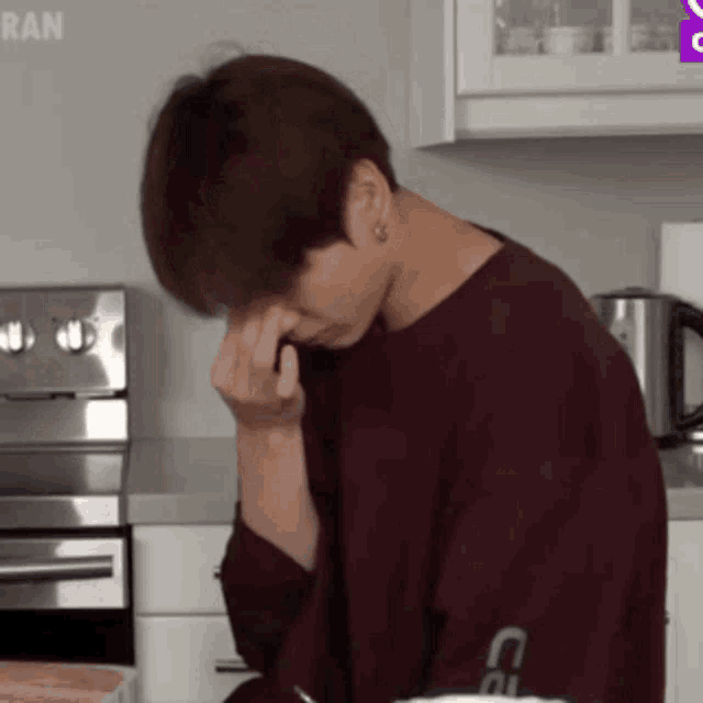 a man in a maroon shirt is sitting in a kitchen with his hand on his face .