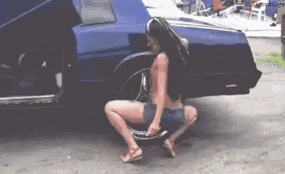 a woman is squatting down in front of a blue car holding a tire .