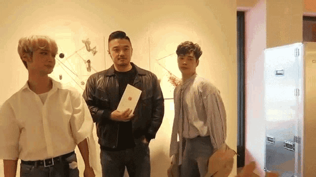 three men are standing next to each other in front of a wall . one of the men is holding a book .