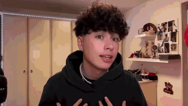 a young man with curly hair is wearing a pearl necklace and a hoodie .