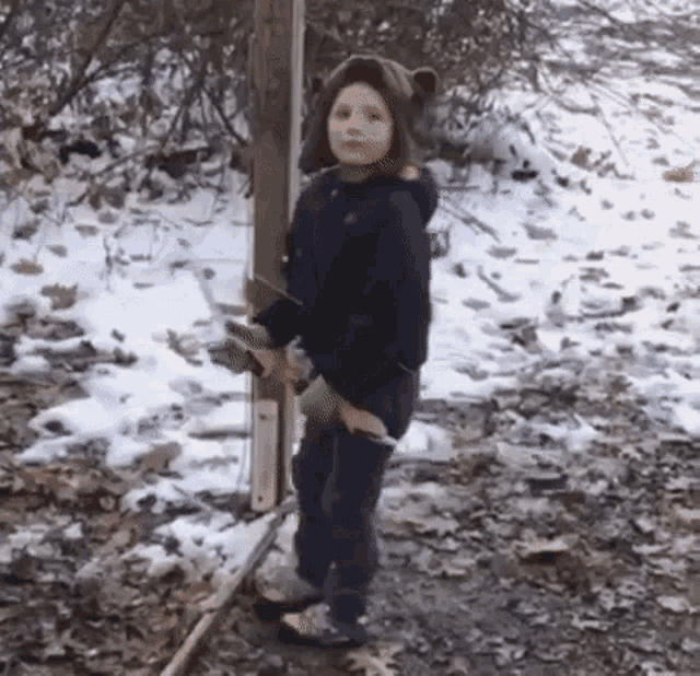 a young boy wearing a bear hat is standing in the snow holding a skateboard .