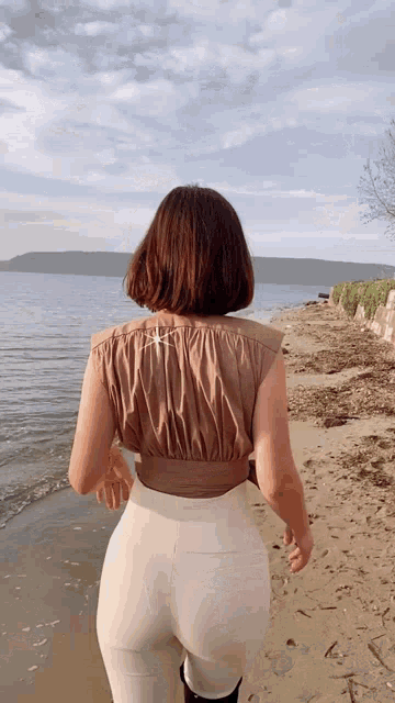 a woman in a brown top and white pants walks on a beach