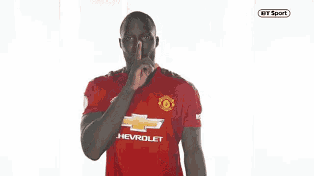a man in a red shirt is pointing at the camera with his finger on his lips .