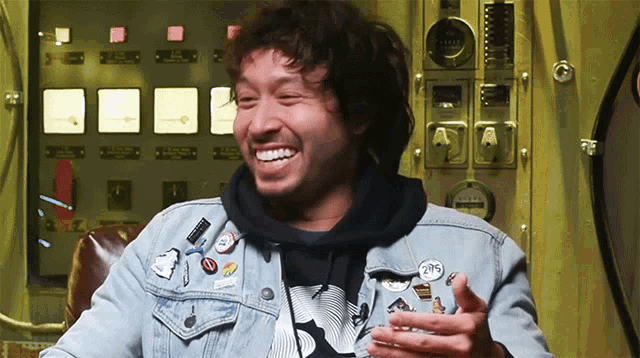 a man wearing a denim jacket and a black hoodie is smiling in front of a control panel