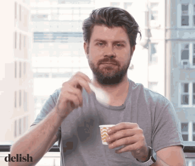 a man with a beard is holding a cup of coffee and the word delish is on the bottom right