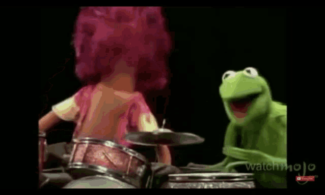 kermit the frog is playing the drums next to a doll with pink hair .