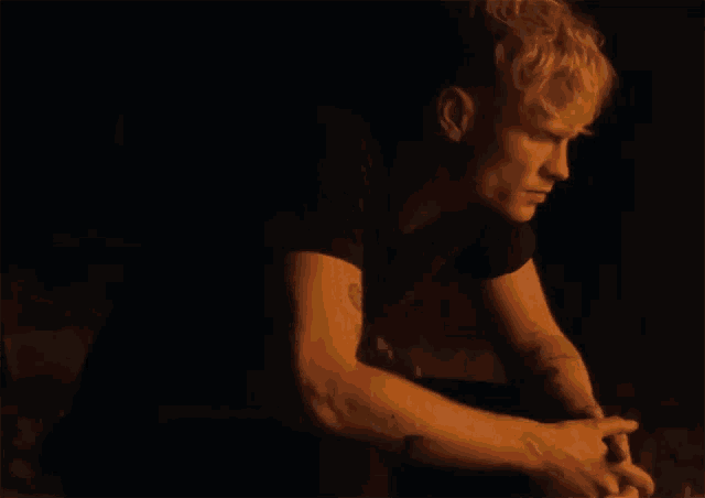 a man with blonde hair and a tattoo on his arm is sitting in the dark