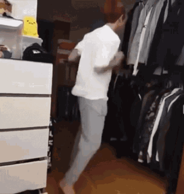 a man in a white shirt is dancing in a room with clothes hanging on the wall .