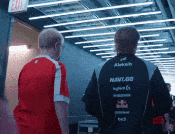 two men are walking down a hallway and one of them is wearing an aleksib navi.gg jacket