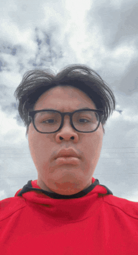 a man wearing glasses and a red shirt looks at the camera with a cloudy sky in the background