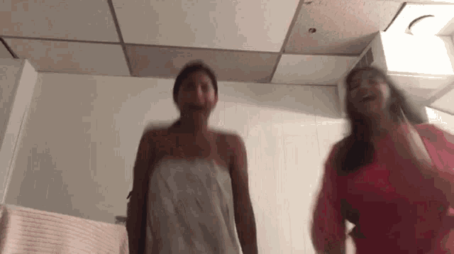 two women are standing next to each other in a bathroom . one of the women is wrapped in a towel .